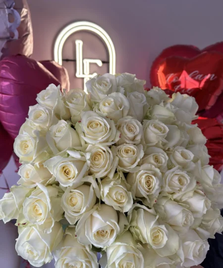 Large bouquet of white roses with elegant long stems, arranged in front of heart-shaped balloons and the Lu La Fleur logo