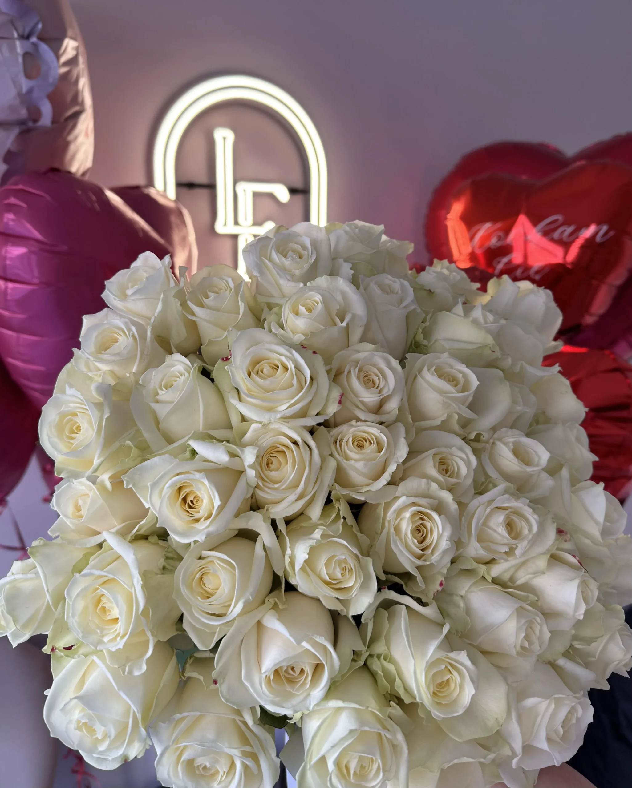 Large bouquet of white roses with elegant long stems, arranged in front of heart-shaped balloons and the Lu La Fleur logo