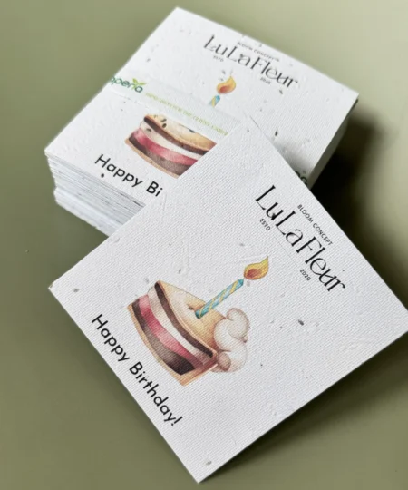 Kartka z okazji urodzin marki „Lu La Fleur” z ilustracją kolorowego kawałka tortu z zapaloną świeczką. Na górze widnieje logo „Lu La Fleur – Bloom Concept ESTD 2020”, a na dole napis „Happy Birthday!”. W tle stos identycznych kartek.