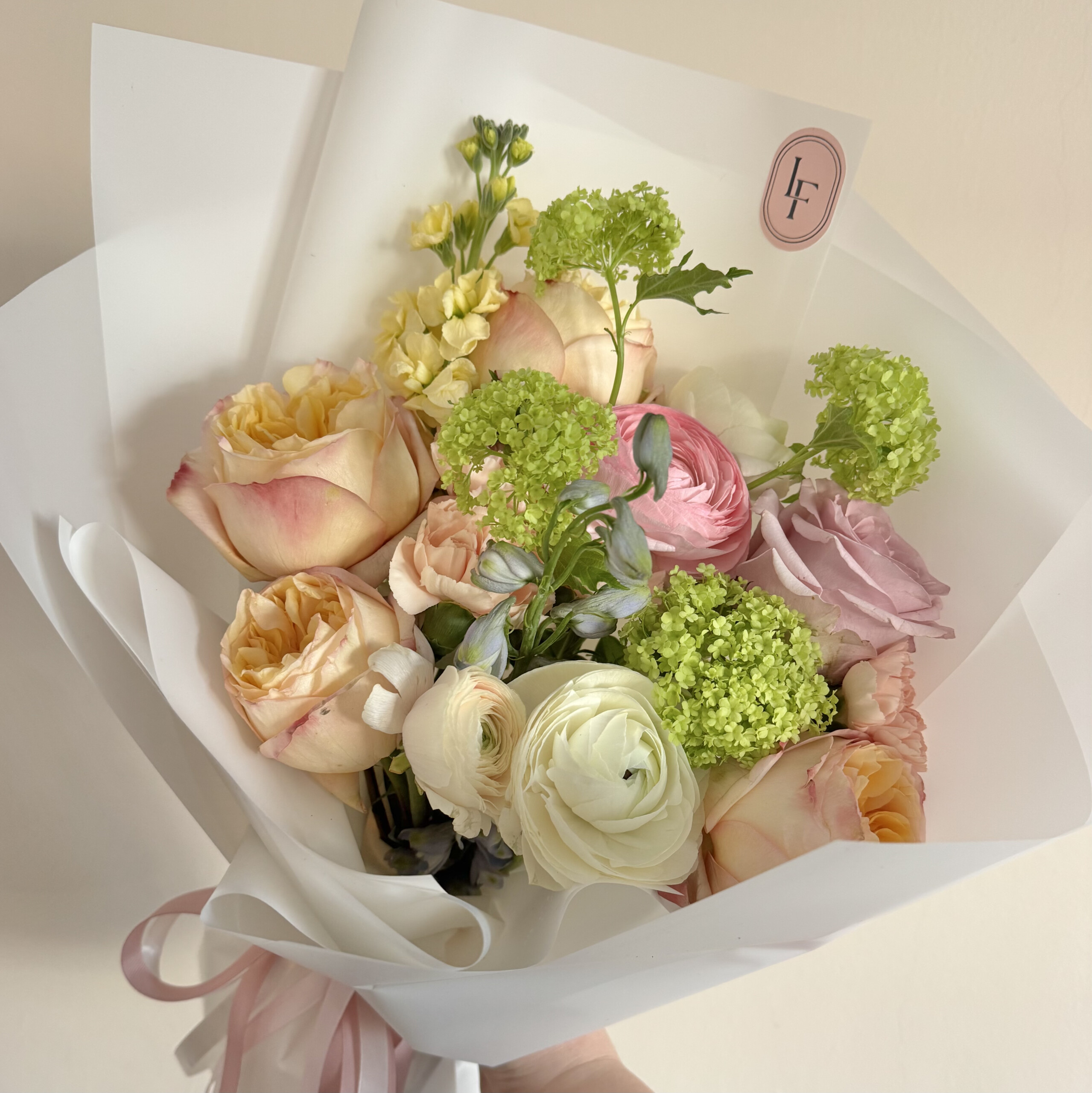 Romantyczny bukiet z pastelowych róż, jaskrów i eustomy z dodatkiem zielonych kwiatów sezonowych, elegancko ułożony przez LuLaFleur.